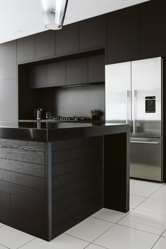 Photograph of the finished black kitchen showing the timber-clad island, black overheads and stainless fridge